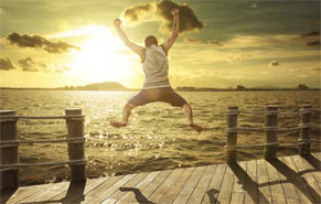 Man in shorts jumping off a short pier with hands hands and legs spread in joy.