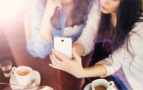 Two women in a coffee shop. One is watching as the other points to the window of her cellphone.